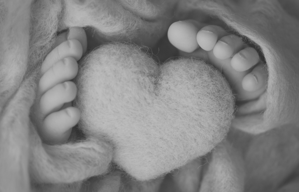 Czarno-białe zdjęcie stóp noworodka otulonych miękkim kocem, z filcowym sercem między stópkami – ilustracja badań genetycznych dziecka po poronieniu.”